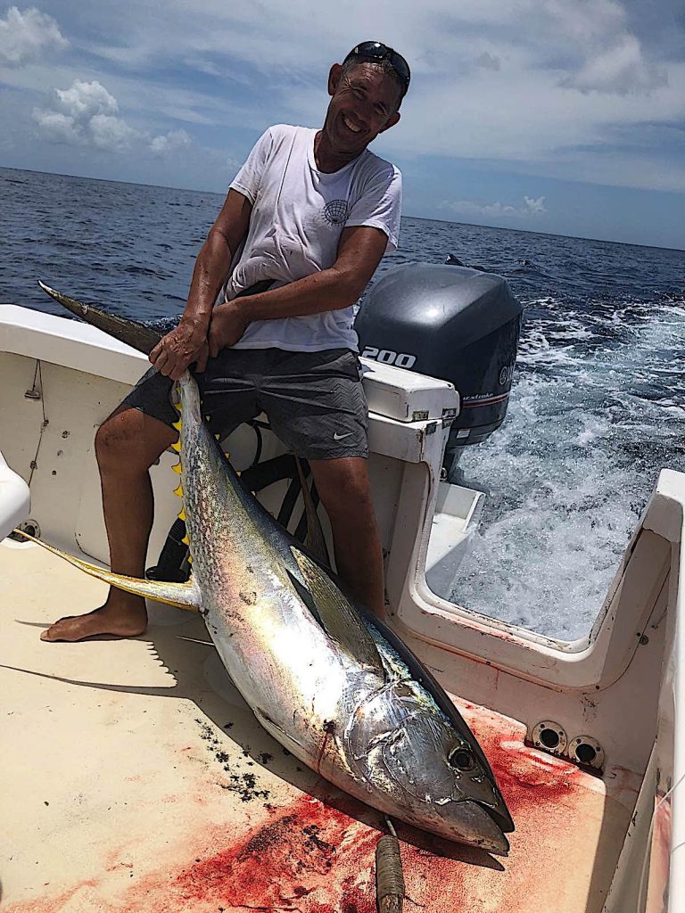 Angler holding a huge yellow fin tuna on the back of the boat. There are blue skies with light cloud.